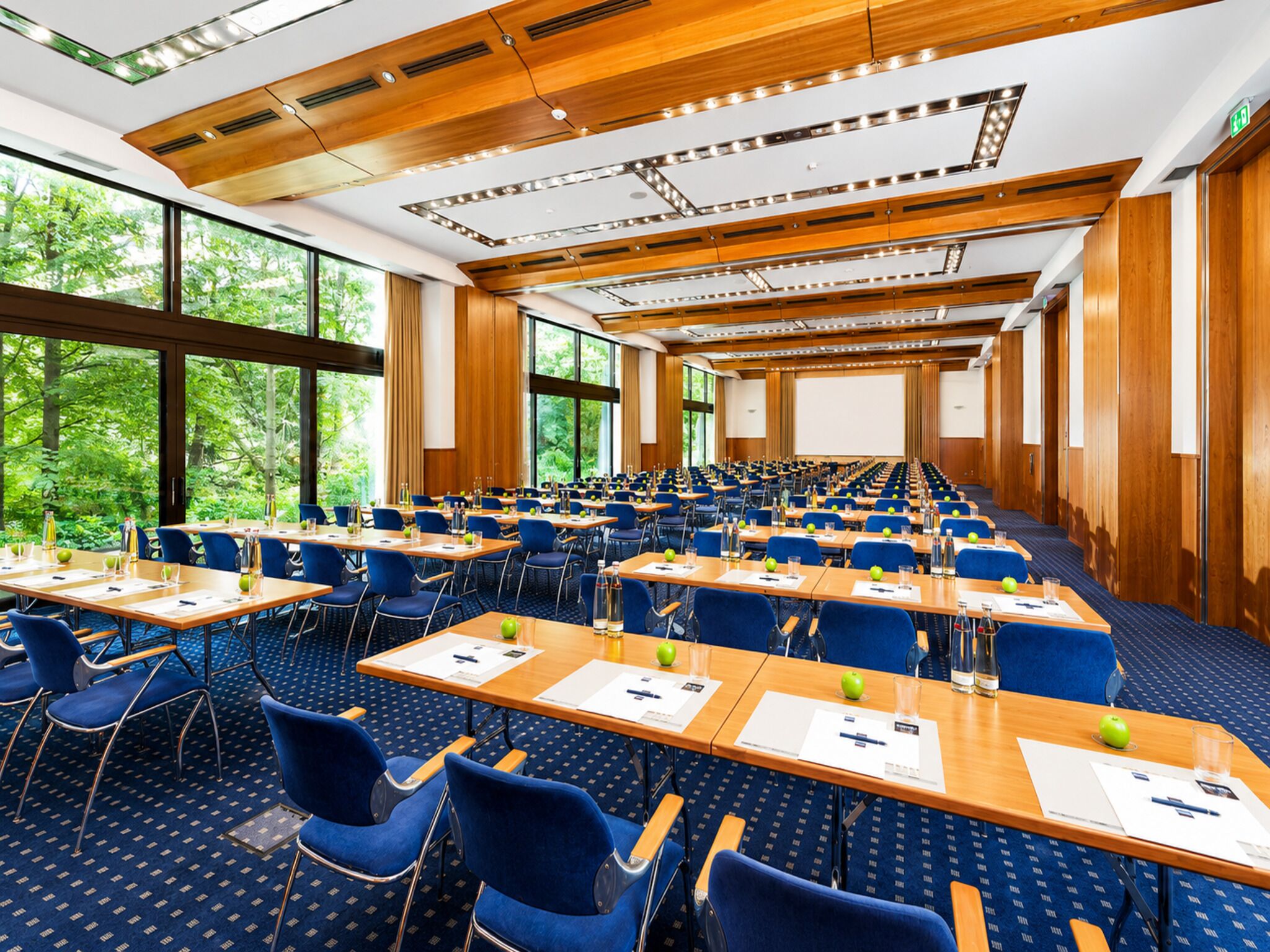 Un salón de conferencias con filas de mesas y sillas, ventanas amplias y vista a zonas verdes.