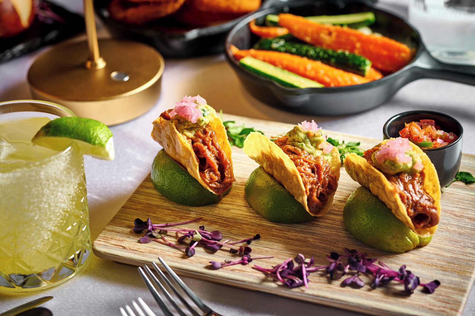 Tacos op limoenhelften met een drankje en een schaal groenten op de achtergrond.