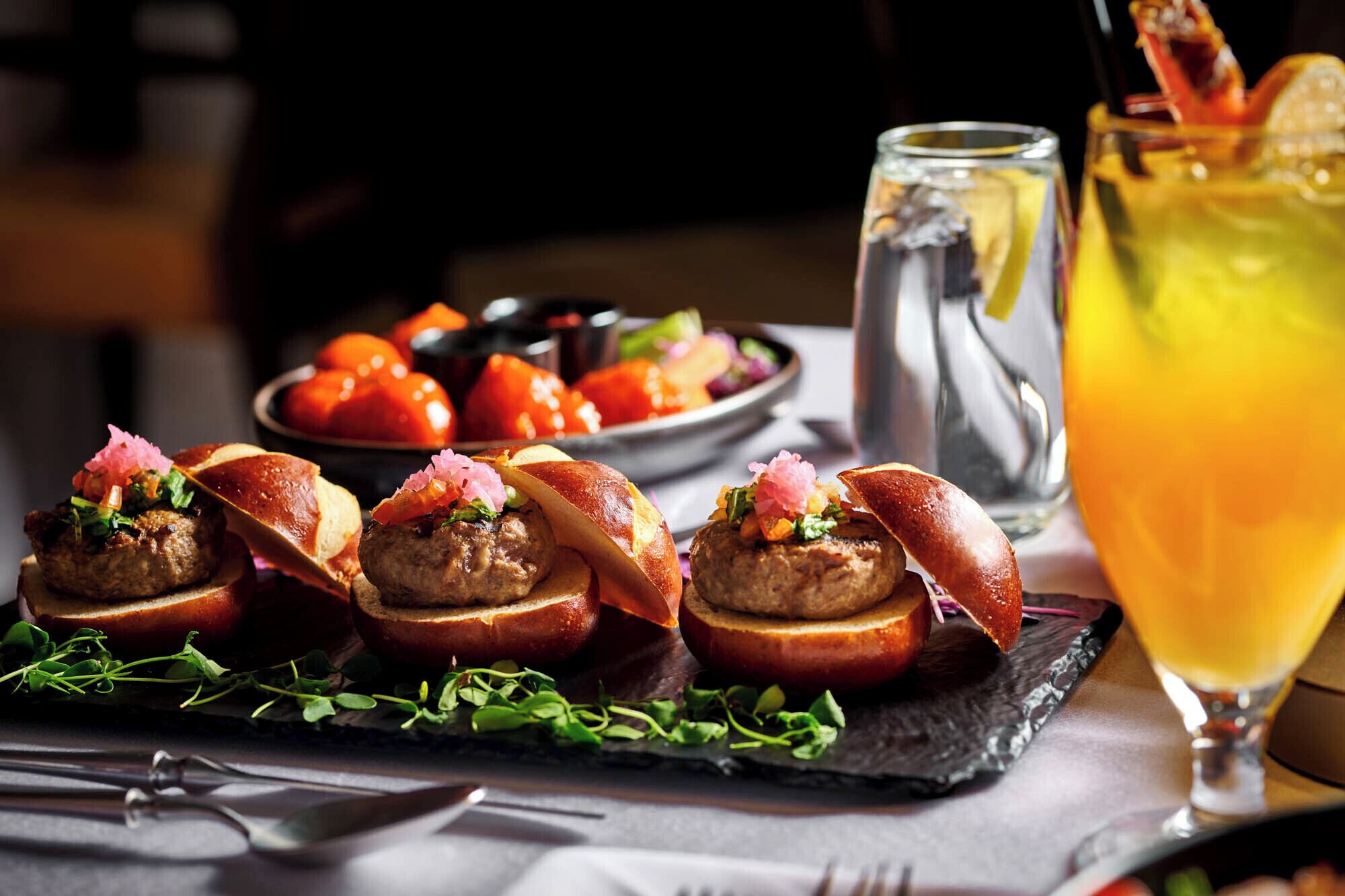 A dining table with a black slate serving platter holding meatballs topped with bacon and garnishes, a pitcher of iced beverage, and a glass of orange juice.