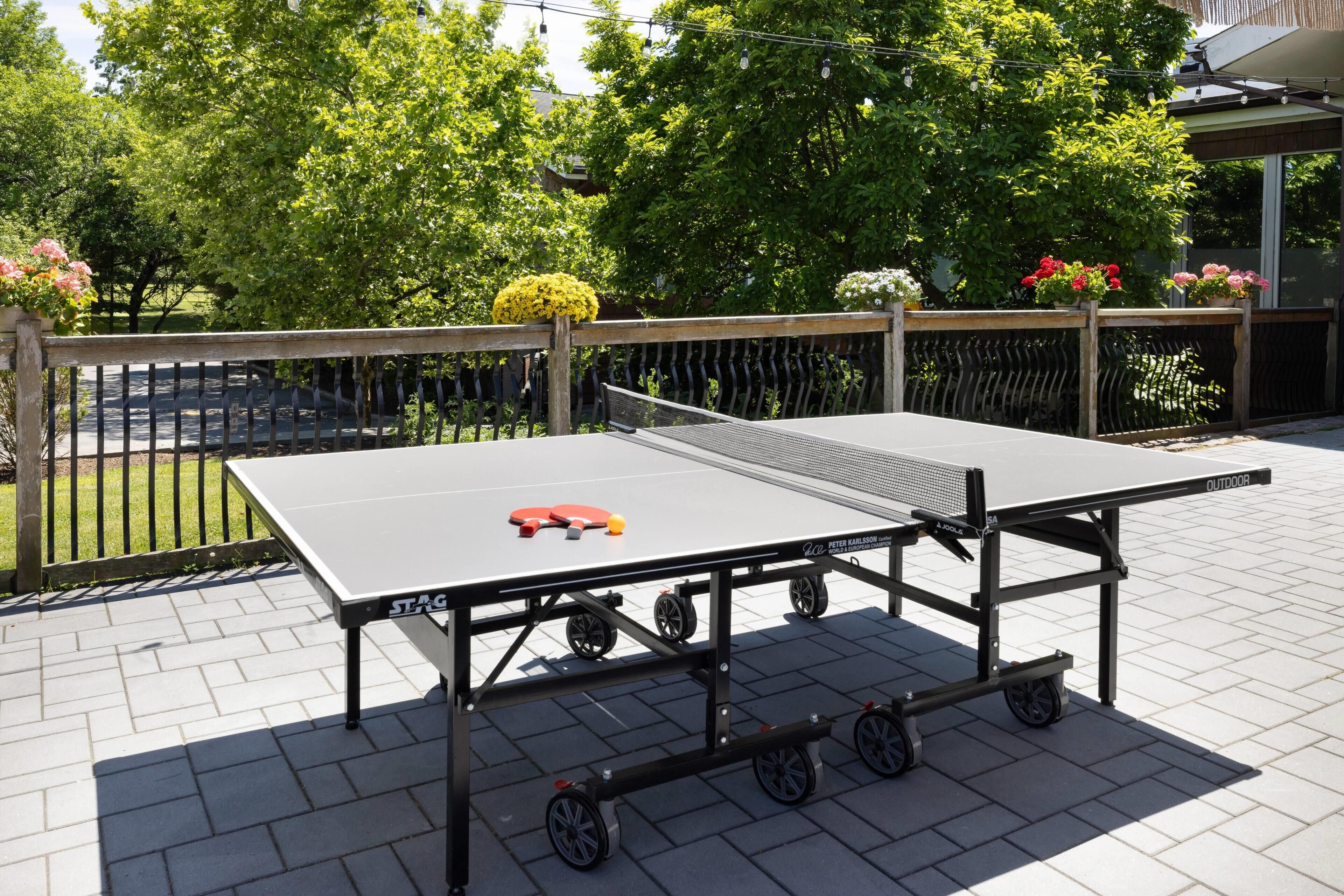 An outdoor ping pong table with wheels, surrounded by a wooden fence and potted plants.