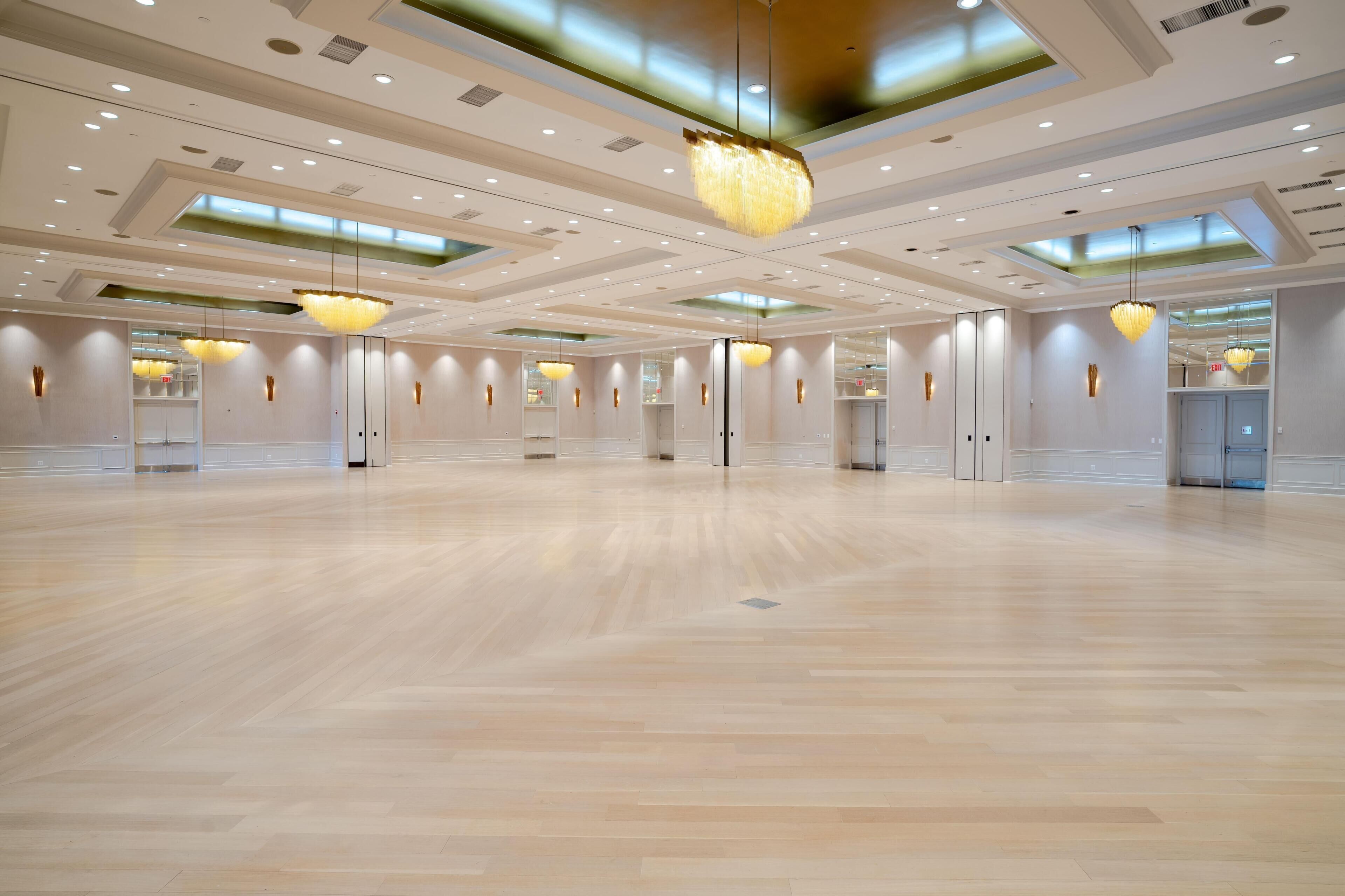 Un salón de eventos amplio en un hotel, con piso de madera lustrosa, iluminación empotrada y candelabros modernos.