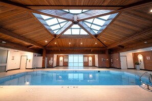 Una piscina interior con techo de madera alto y tragaluces, rodeada de paredes de ladrillo y equipada con señales de seguridad.
