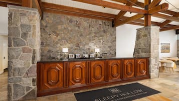 A reception area with a stone wall, wooden reception desk, and a black doormat with the hotel name.