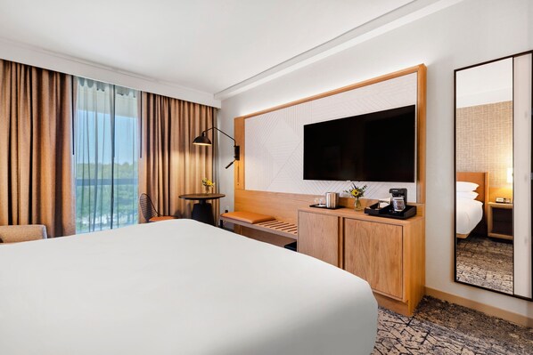 Habitación de hotel con una cama grande, un escritorio con cafetera, un televisor y un espejo amplio.