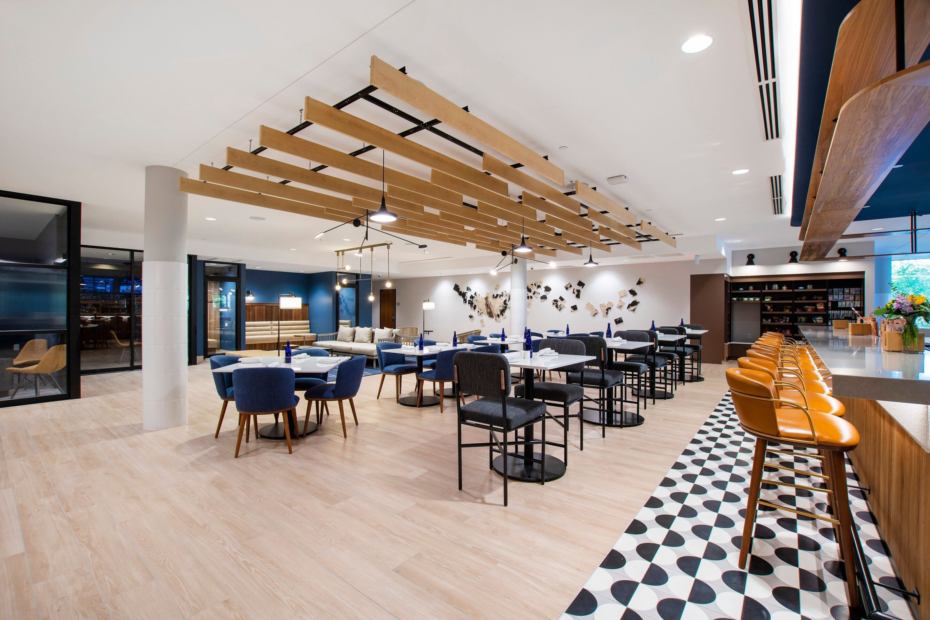 A modern restaurant with wooden ceiling beams, a checkered floor, and a long bar with orange stools.
