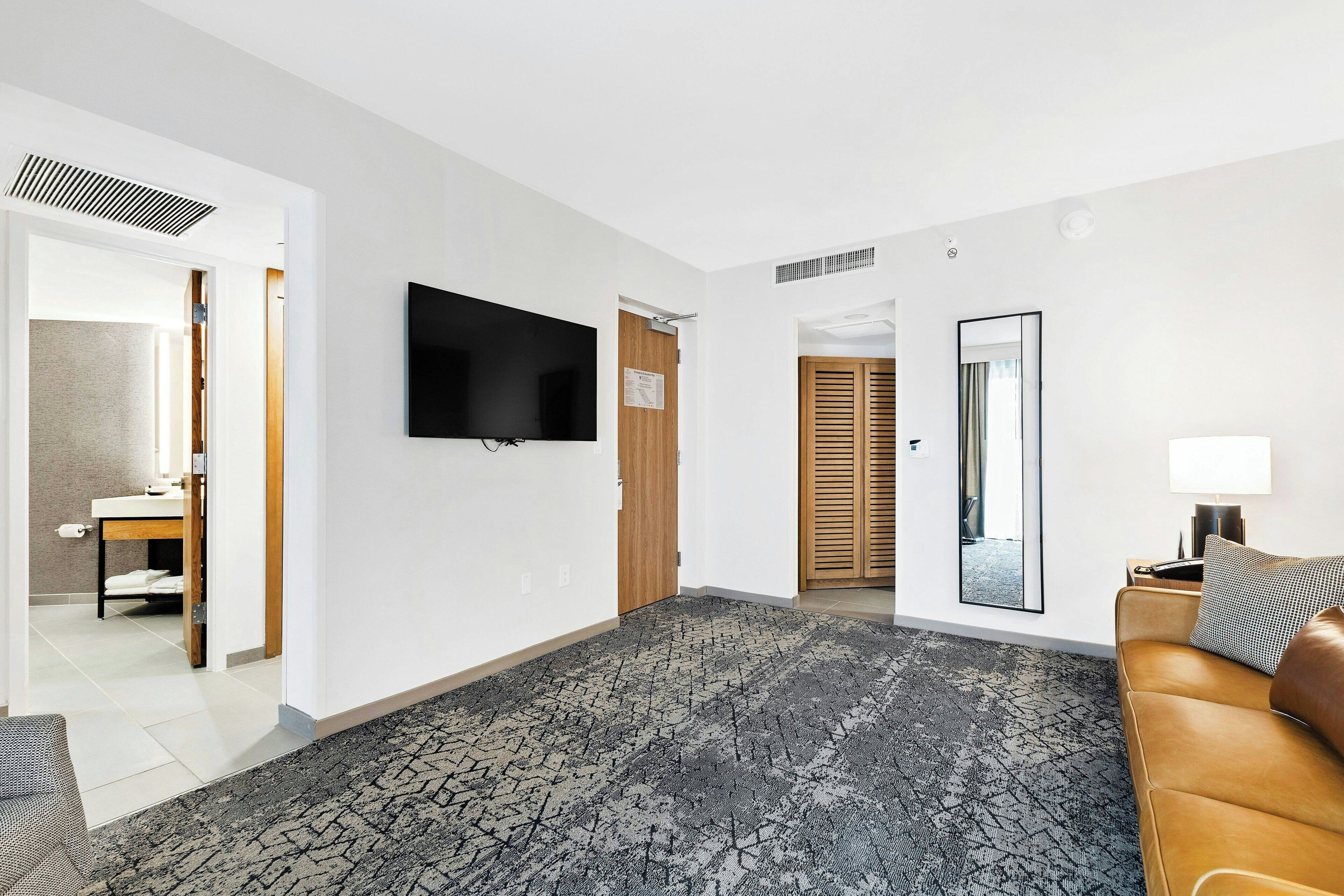 A hotel room with a patterned carpet, a flat-screen TV, a wooden door, a mirror, a sofa, and a nightstand with a lamp.