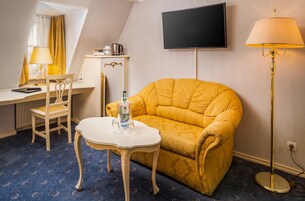 Quarto de hotel com sofá amarelo, uma pequena mesa branca, escrivaninha com cadeira, televisão de tela plana e abajur.