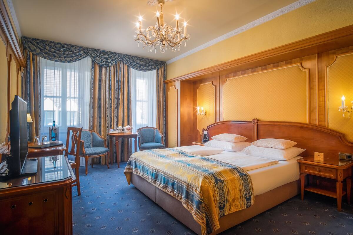Habitación de hotel con una cama grande, un escritorio con televisor, una silla y una araña.