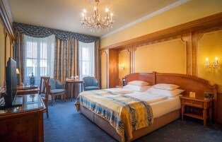 Quarto de hotel com uma cama grande, uma escrivaninha com televisão, uma cadeira e um candelabro.