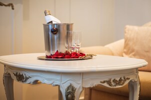 Uma mesa branca com um balde de prata contendo uma garrafa e dois copos, rodeada por pétalas de rosa vermelhas.