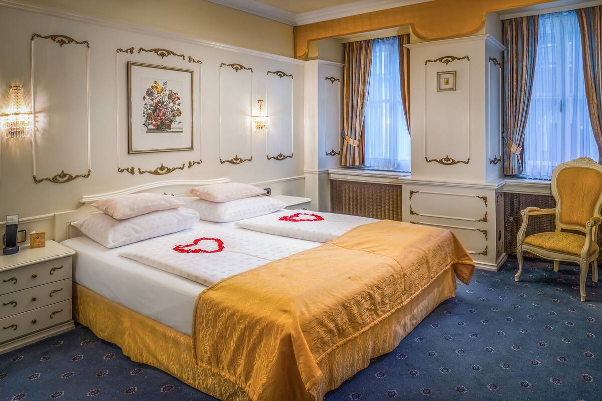 Una habitación de hotel con una cama grande, dos mesitas de noche, una silla, una ventana con cortinas y un cuadro colgado en la pared.