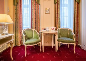 Quarto com duas poltronas estofadas em verde, uma mesinha redonda com um bule e xícaras, um abajur e um chão coberto por um tapete vermelho.