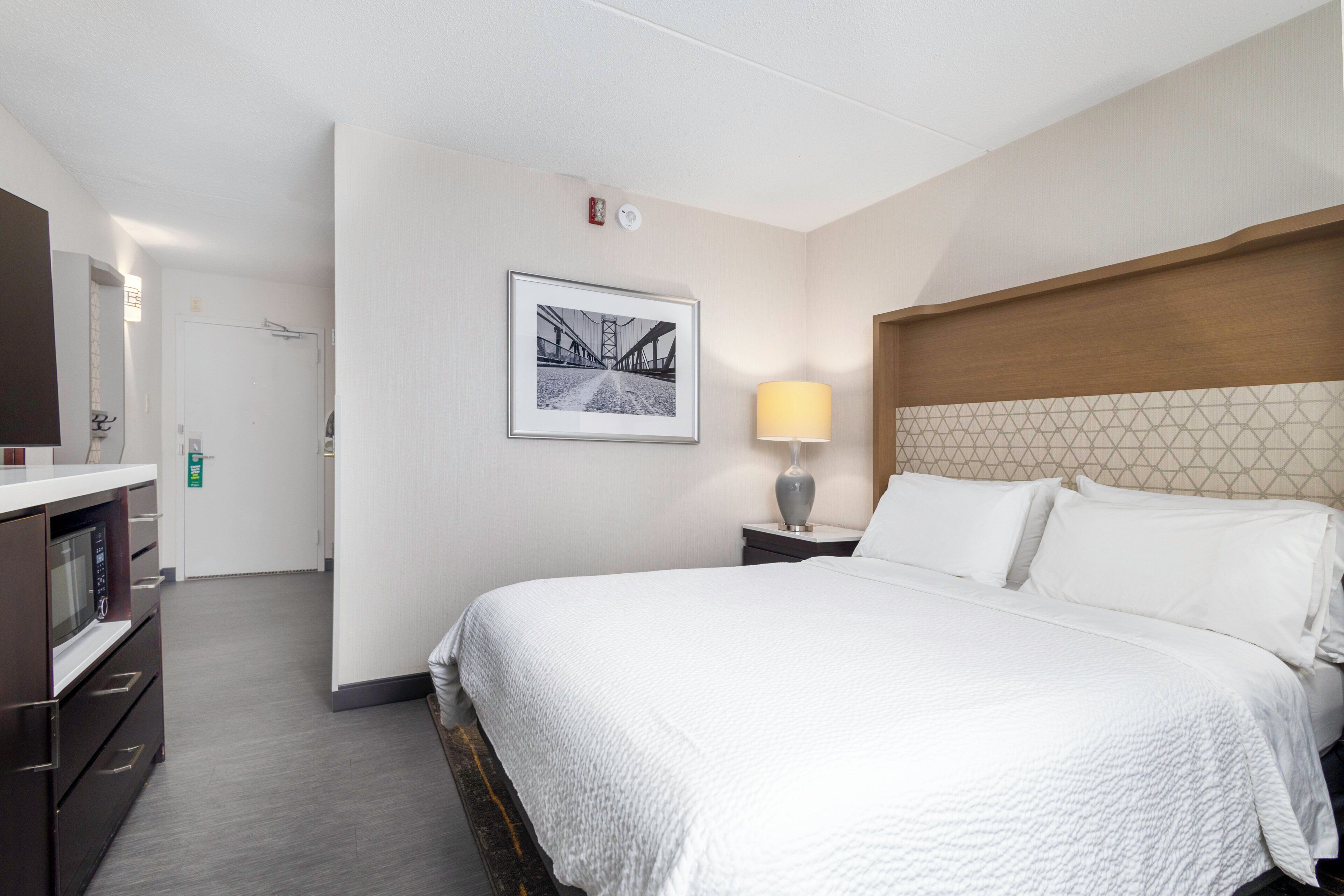 A hotel room with a bed, a bedside lamp, a framed picture on the wall, and a microwave in the corner.