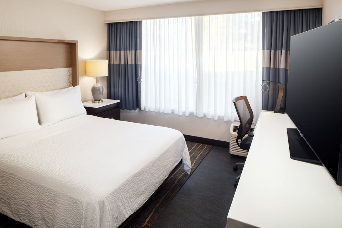 A hotel room with a large bed, a desk with a chair, a TV, a lamp, and a window with curtains.