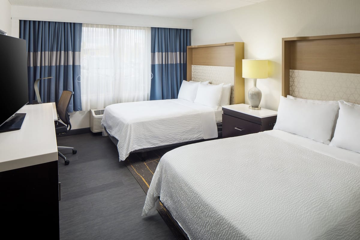 A hotel room with two beds, a desk, a chair, a TV, and a lamp.