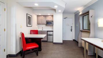 A compact hotel room with a kitchenette, a dining area with a red chair, and a small table.