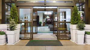 A hotel entrance with double doors, potted plants, and a visible interior including a reception area and a staircase. - Holiday Inn Weirton - Steubenville Area by IHG (Weirton)