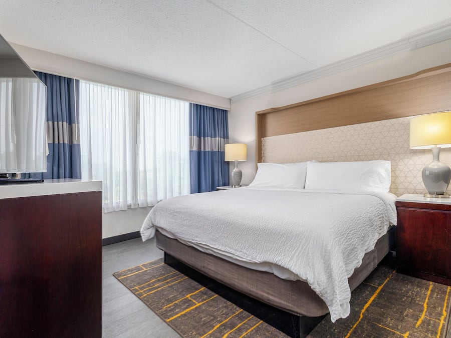 Habitación de hotel con cama, escritorio con televisor, mesita de noche con lámpara y ventana con cortinas.