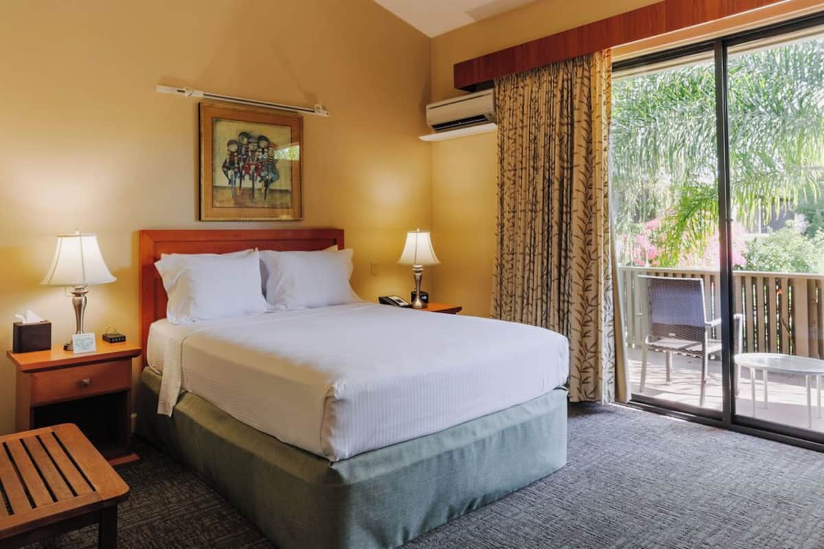 A hotel room with a large bed, two bedside tables with lamps, a painting on the wall, and a balcony with a view of greenery.