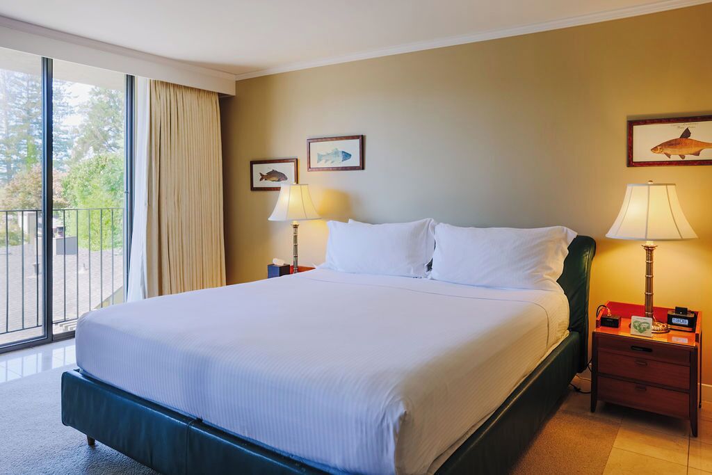 A hotel room with a large bed, two bedside tables with lamps, a window with curtains, and framed pictures on the wall.