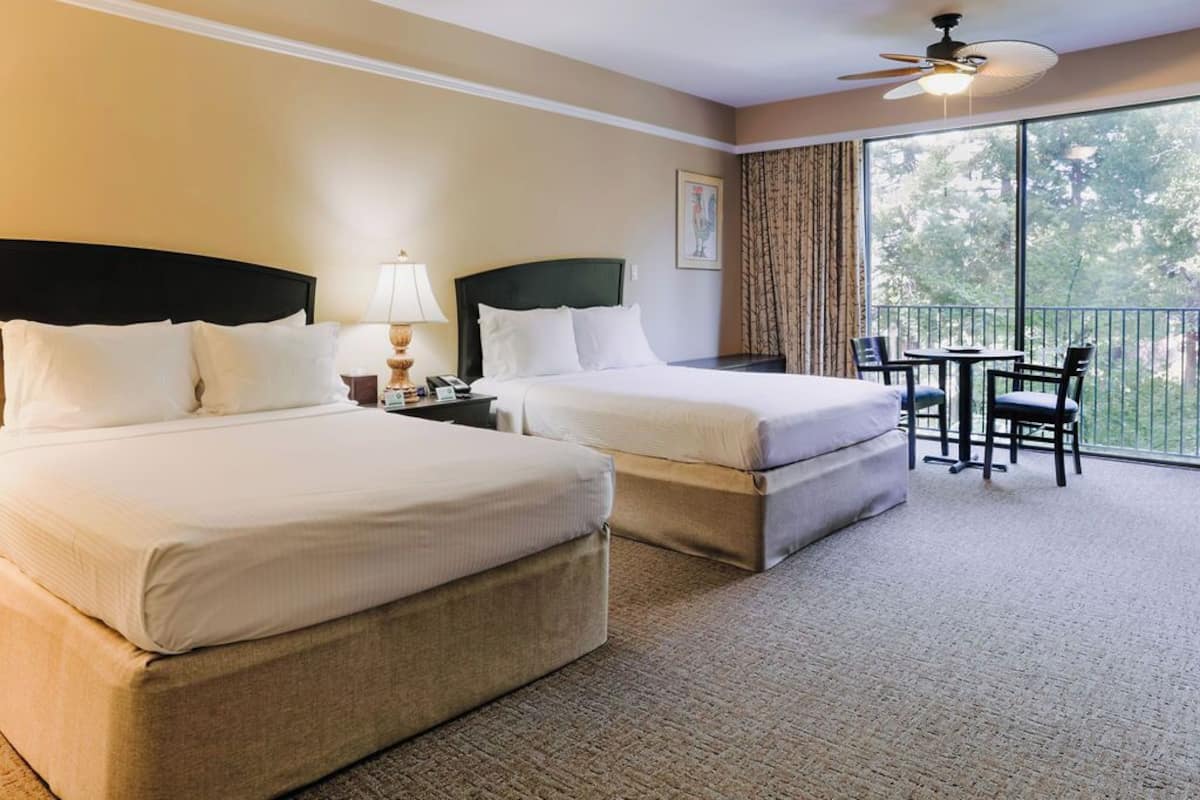 A hotel room with two beds, a balcony with a table and chairs, and a large window offering a view of trees.