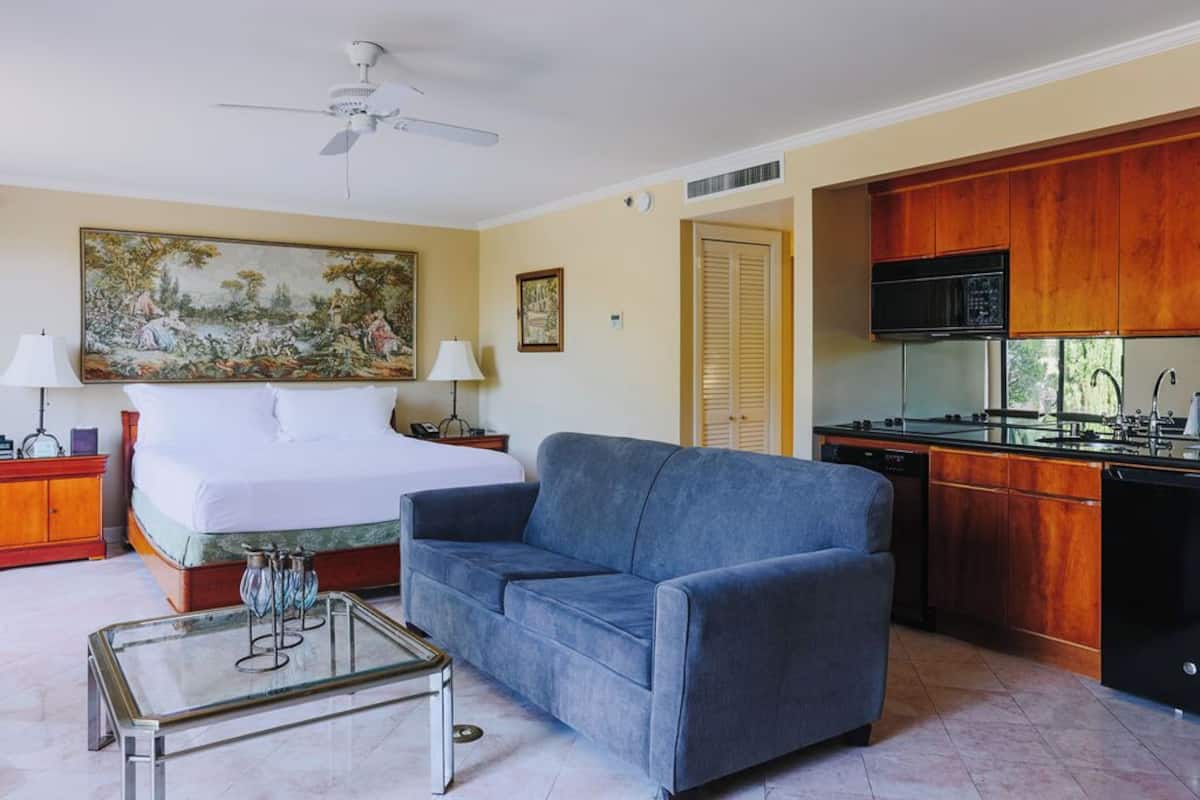 A bedroom with a bed, a blue sofa, a glass coffee table, a ceiling fan, and a kitchen area with wooden cabinets and appliances.