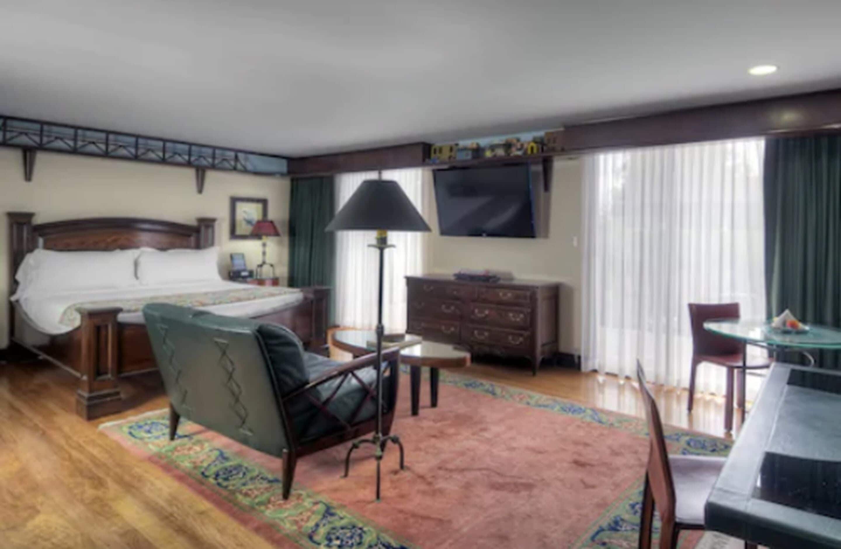 A hotel room with a bed, a desk, a chair, a dresser, a lamp, and a TV.