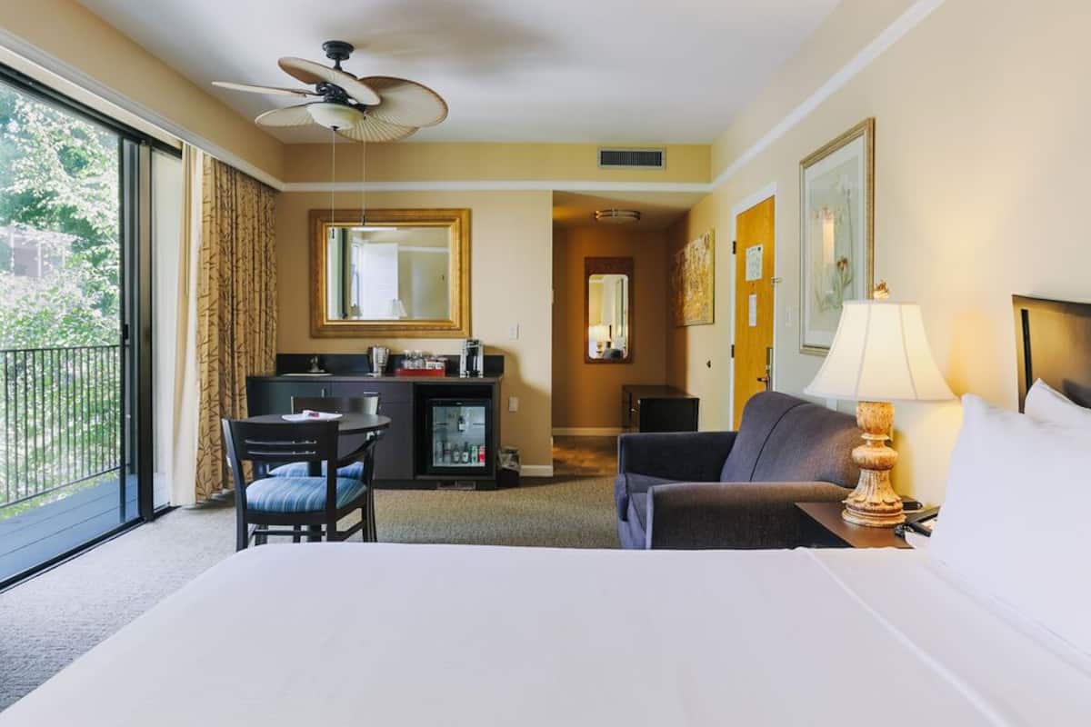 A hotel room with a bed, a desk, a chair, a minibar, a ceiling fan, and a large window.