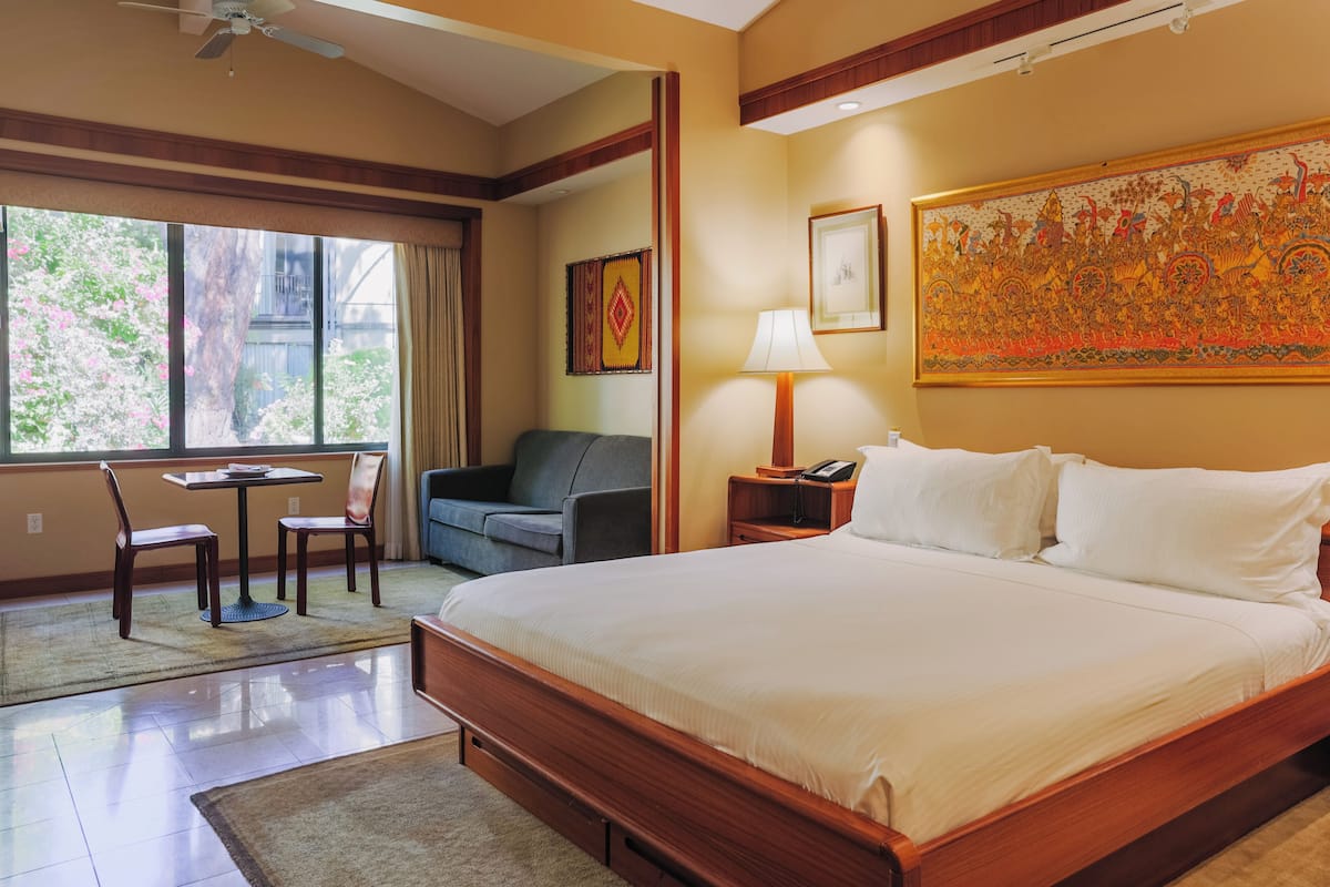 A hotel room with a large bed, a desk with a chair, a sofa, a nightstand with a lamp, and a large painting on the wall.