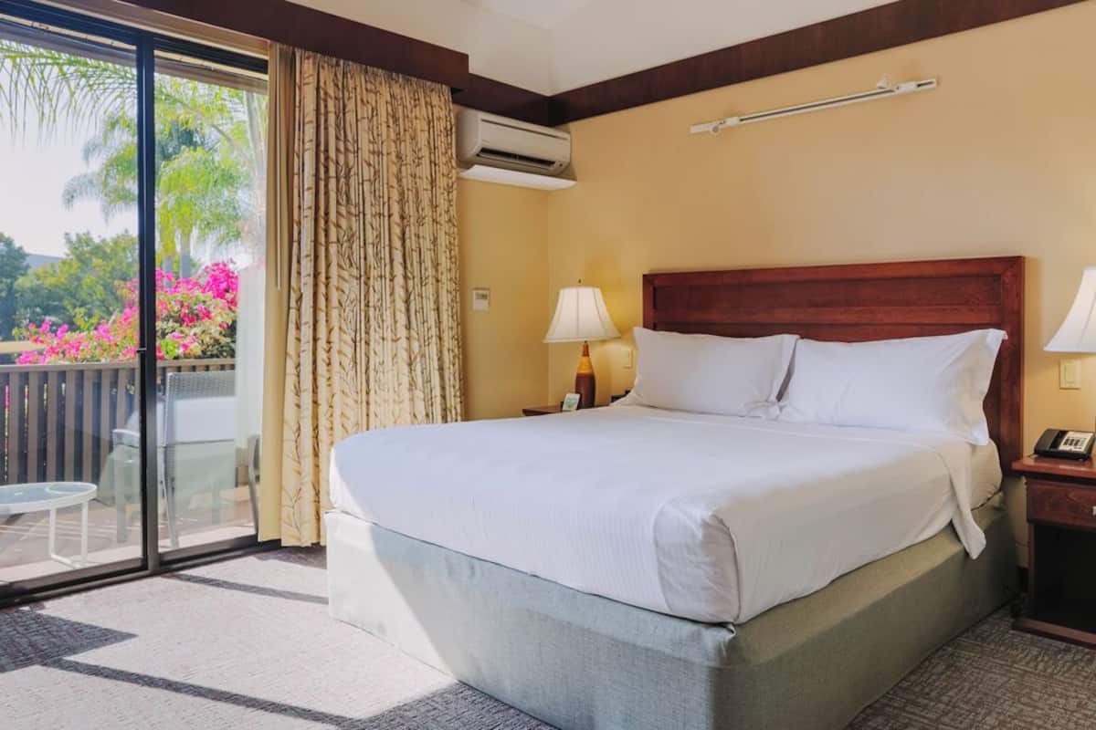 A hotel room with a large bed, a balcony with a view of greenery and flowers, a sliding glass door, and a small table with chairs.