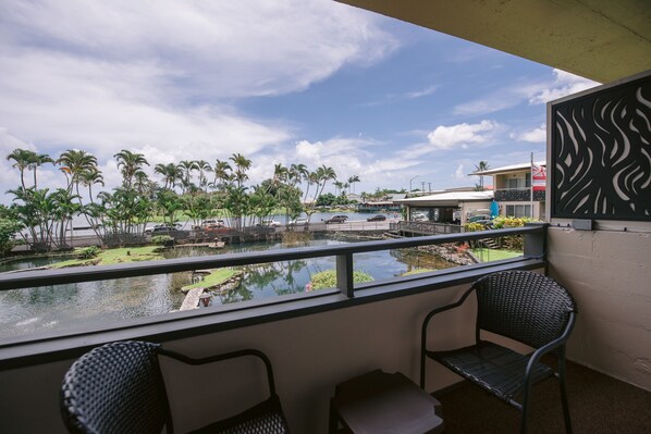 A balcony with two chairs, a view of a water body, palm trees, and a building with a flag. - SCP Hilo Hotel (Hilo)