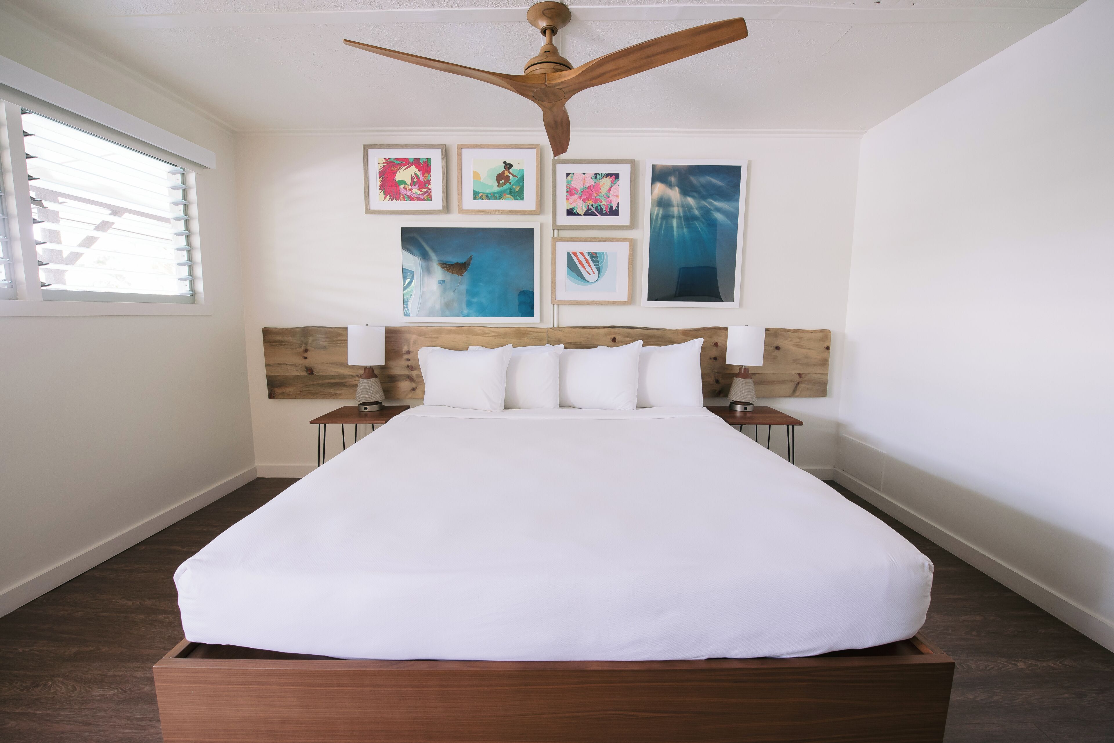 A bedroom with a wooden bed frame, white bedding, a ceiling fan, and four framed artworks on the wall.