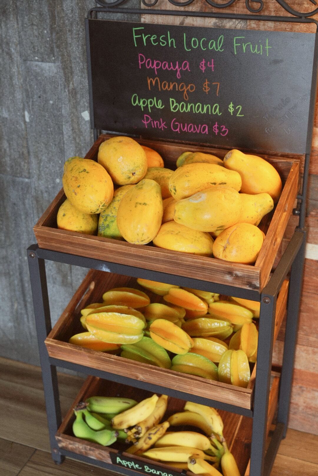 A wooden fruit stand with a chalkboard sign listing prices for papaya, mango, apple banana, and pink guava.