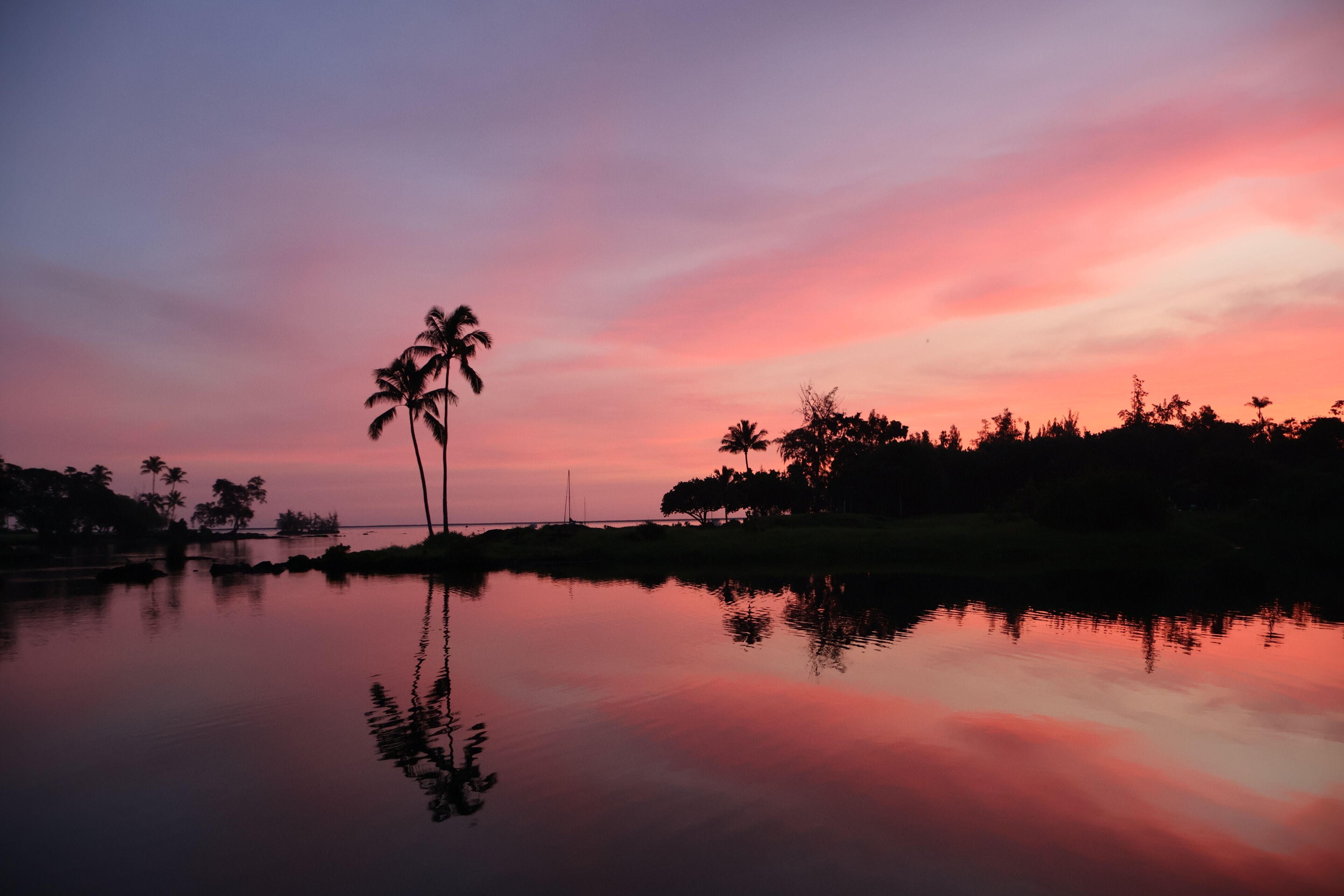 A calm body of water reflecting a sunset sky with palm trees on the horizon.