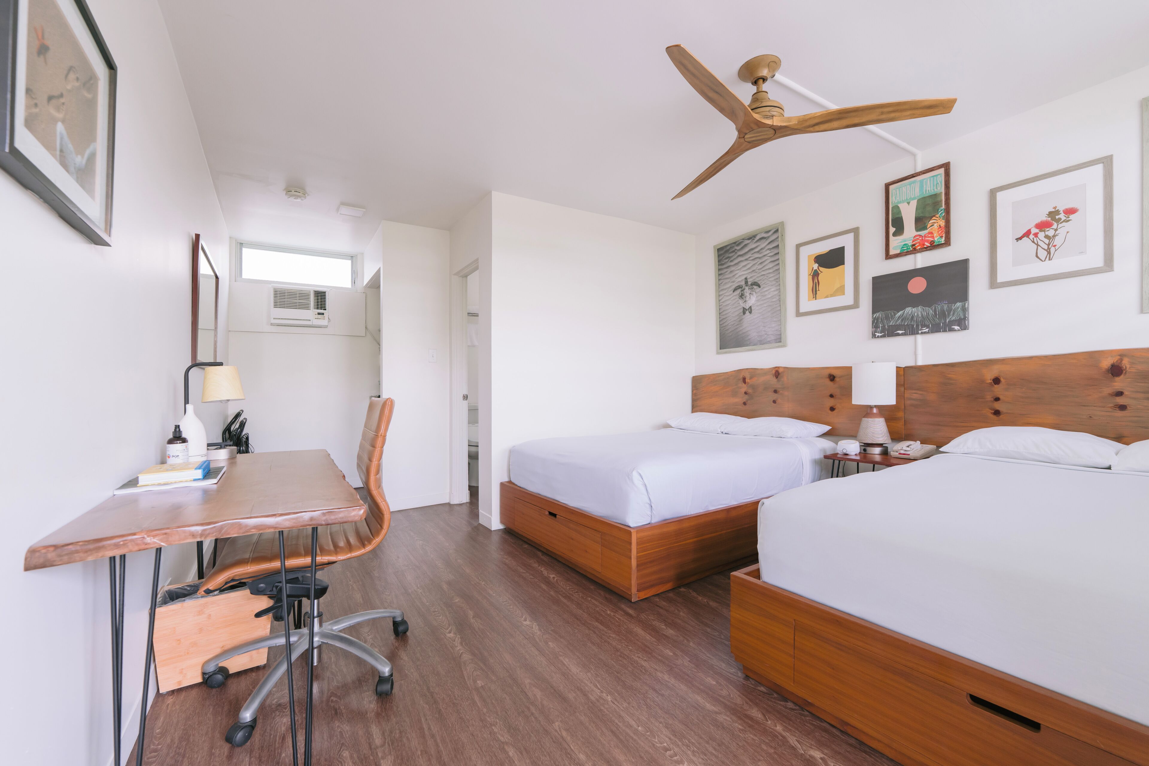 Habitació d'hotel amb dos llits, un escriptori, una cadira, ventilador de sostre i obra d'art a les parets.