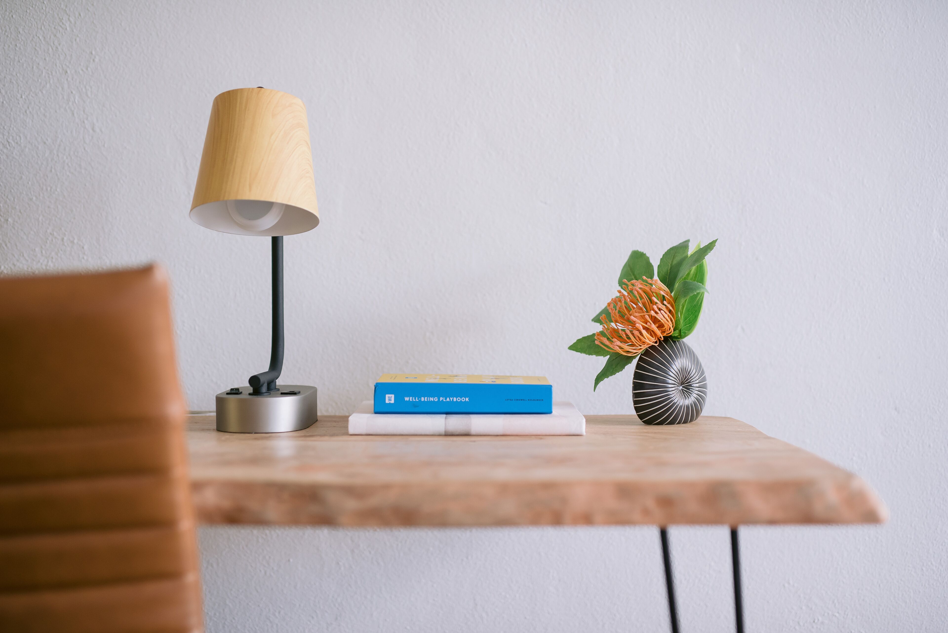 Una mesa de madera con una lámpara, un libro titulado “WELL-BEING PLAYBOOK” y un jarrón decorativo con forma de piña y hojas verdes.