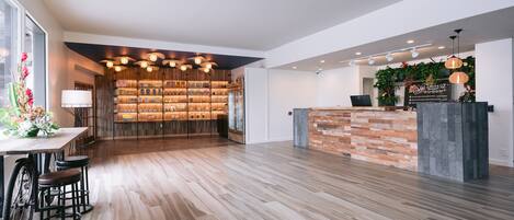 A spacious lobby with a wooden floor, a reception desk with a brick accent wall, and a bar area with shelves of bottles.