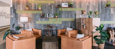 A modern lobby with leather sofas, a coffee table, and a wall decorated with wooden panels and green patterned shelves holding plants and framed pictures.