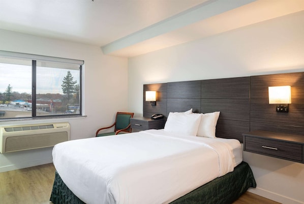 A hotel room with a large bed, a desk with a chair, a window with a view of the outside, and a wall-mounted lamp. - Baymont by Wyndham Bellingham (Bellingham)