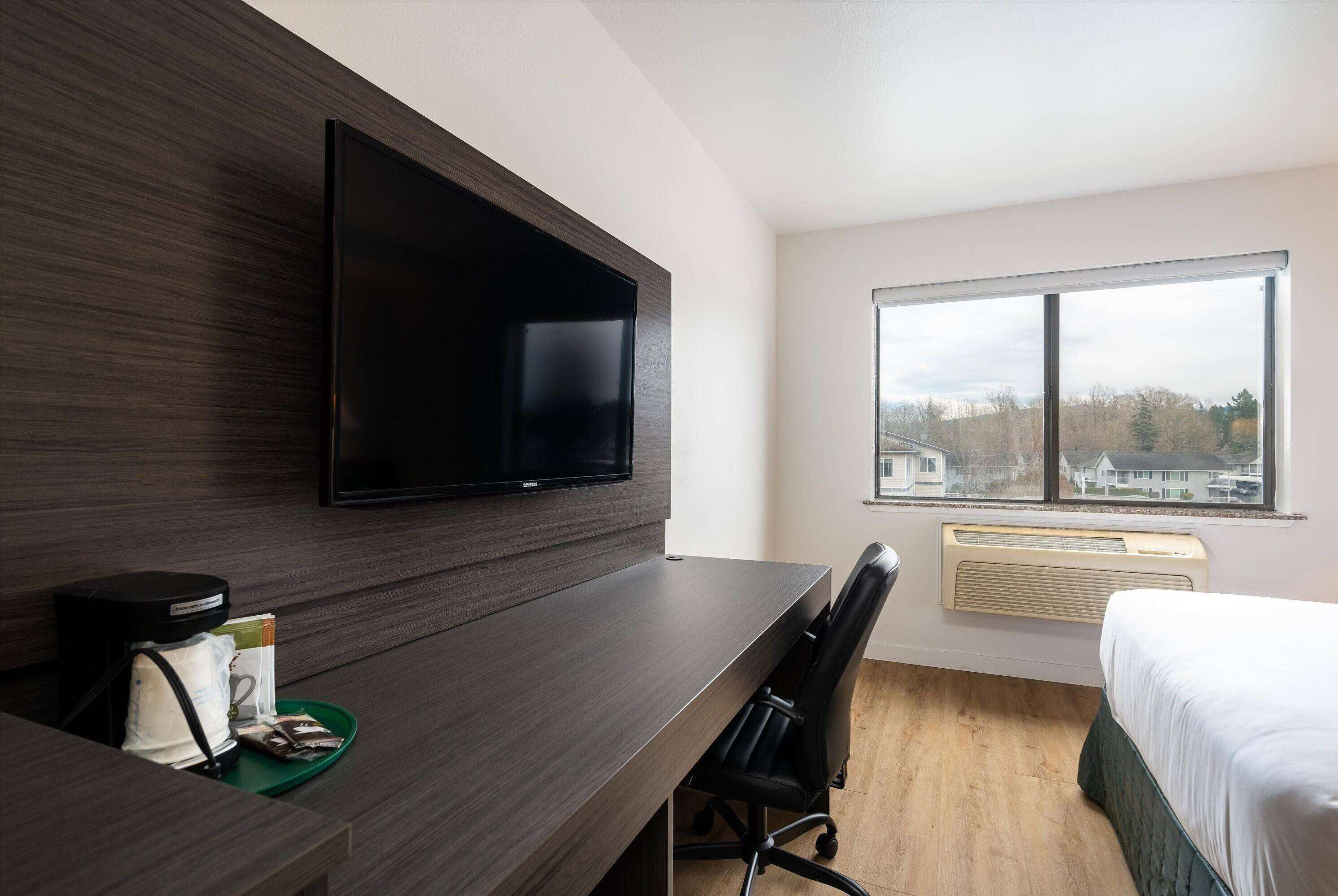 A modern hotel room with a flat-screen TV mounted on the wall, a desk with a chair, a bed, and a large window offering a view of buildings and trees.