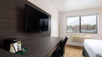 A modern hotel room with a flat-screen TV mounted on the wall, a desk with a chair, a bed, and a large window offering a view of buildings and trees.
