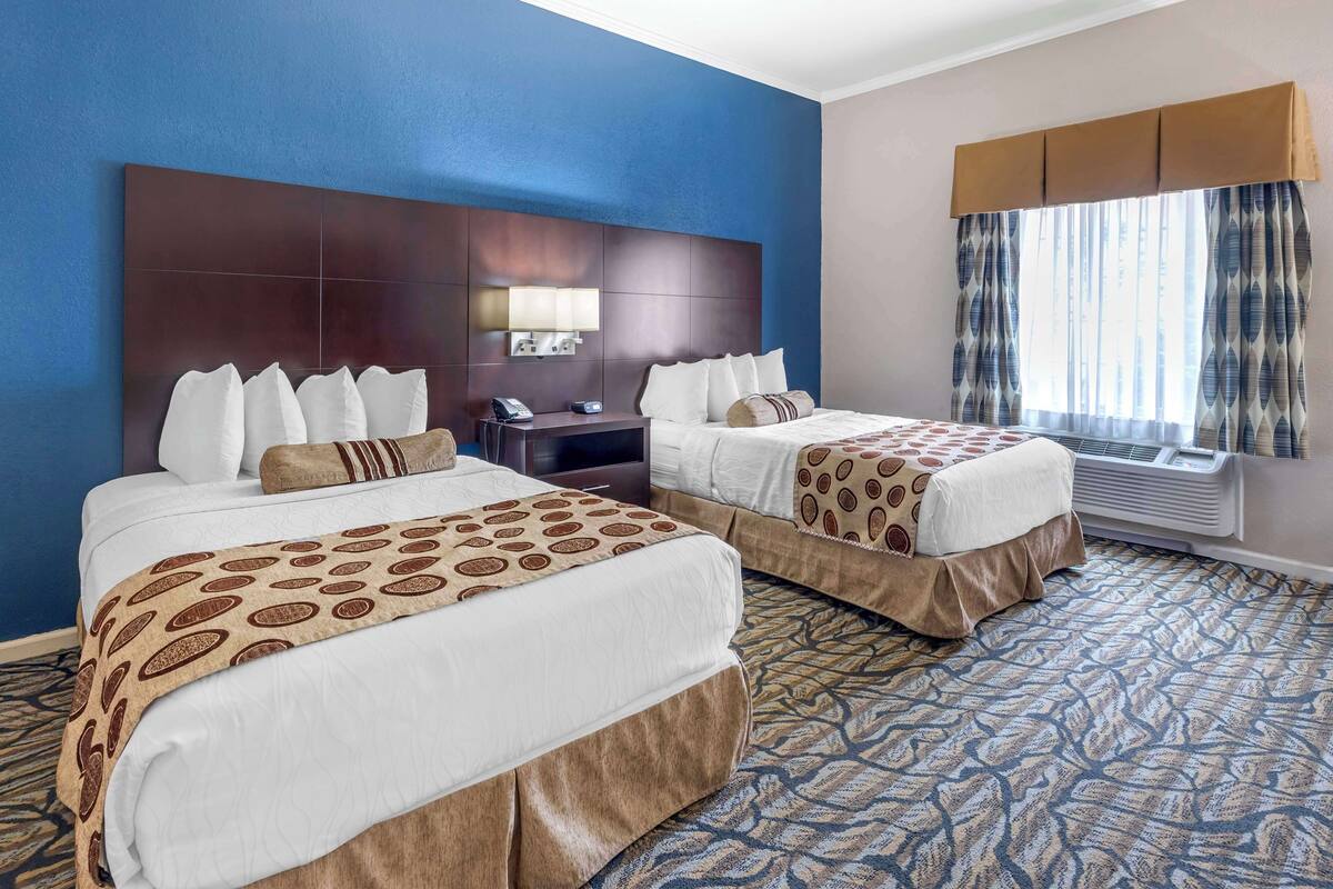 A hotel room with two beds, a large headboard, a bedside table with a lamp, and a window with curtains.