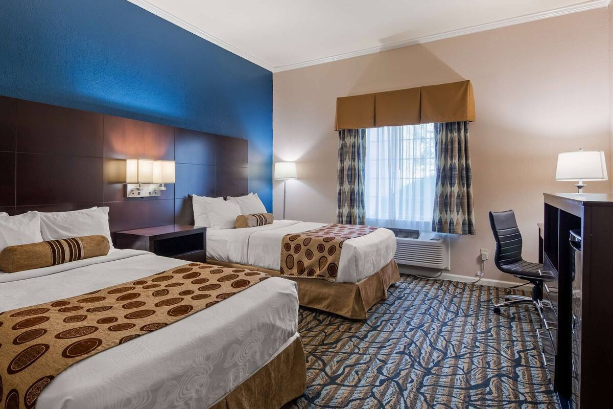 A hotel room with two beds, a desk, a chair, a lamp, and a window with curtains.