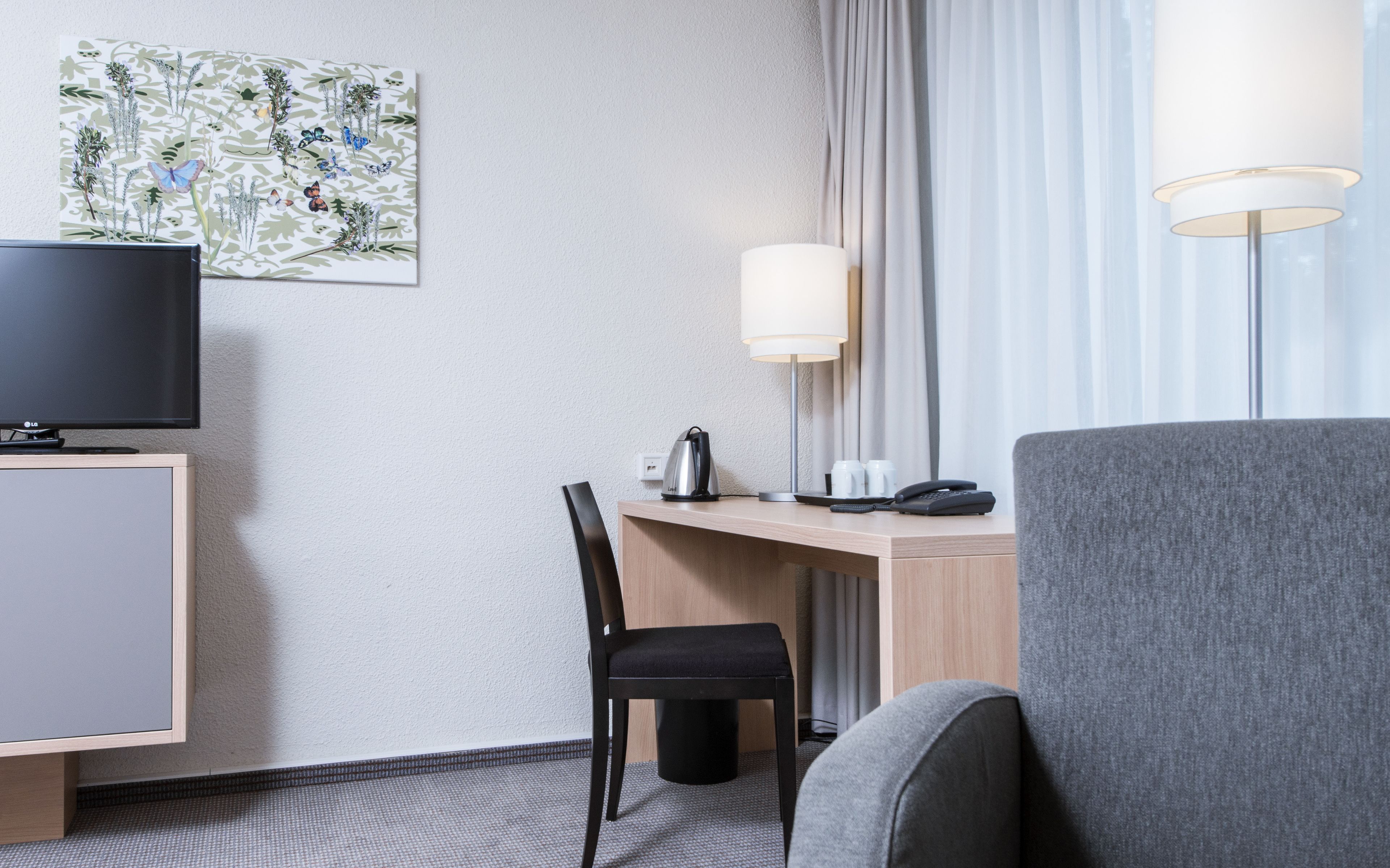 A hotel room with a flat-screen TV, a desk with a lamp, a chair, and a large armchair.