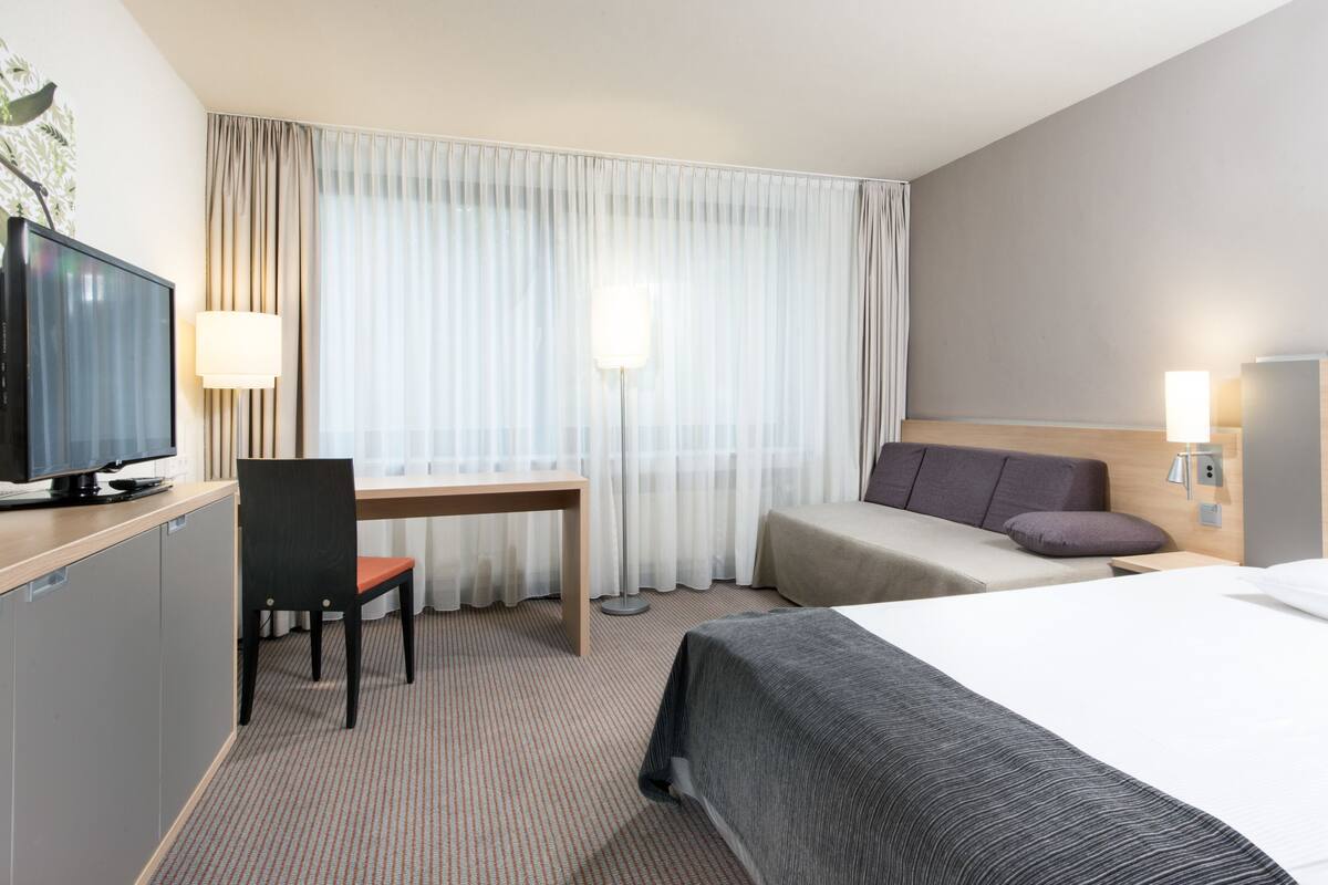 A hotel room with a bed, a desk, a chair, a television, and a window with curtains.