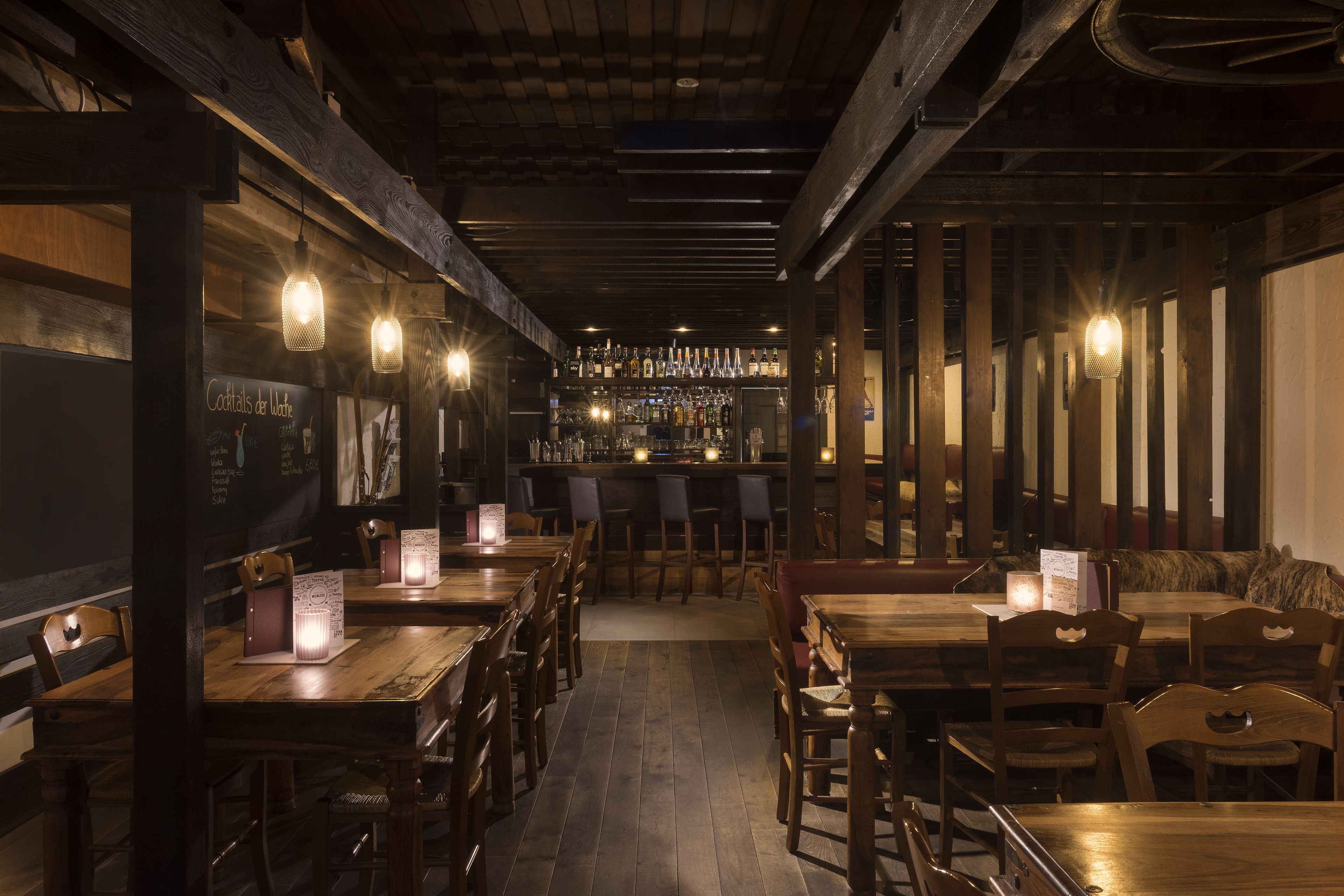 A dimly lit bar area with wooden tables and chairs, a bar counter with bottles, and a menu board.
