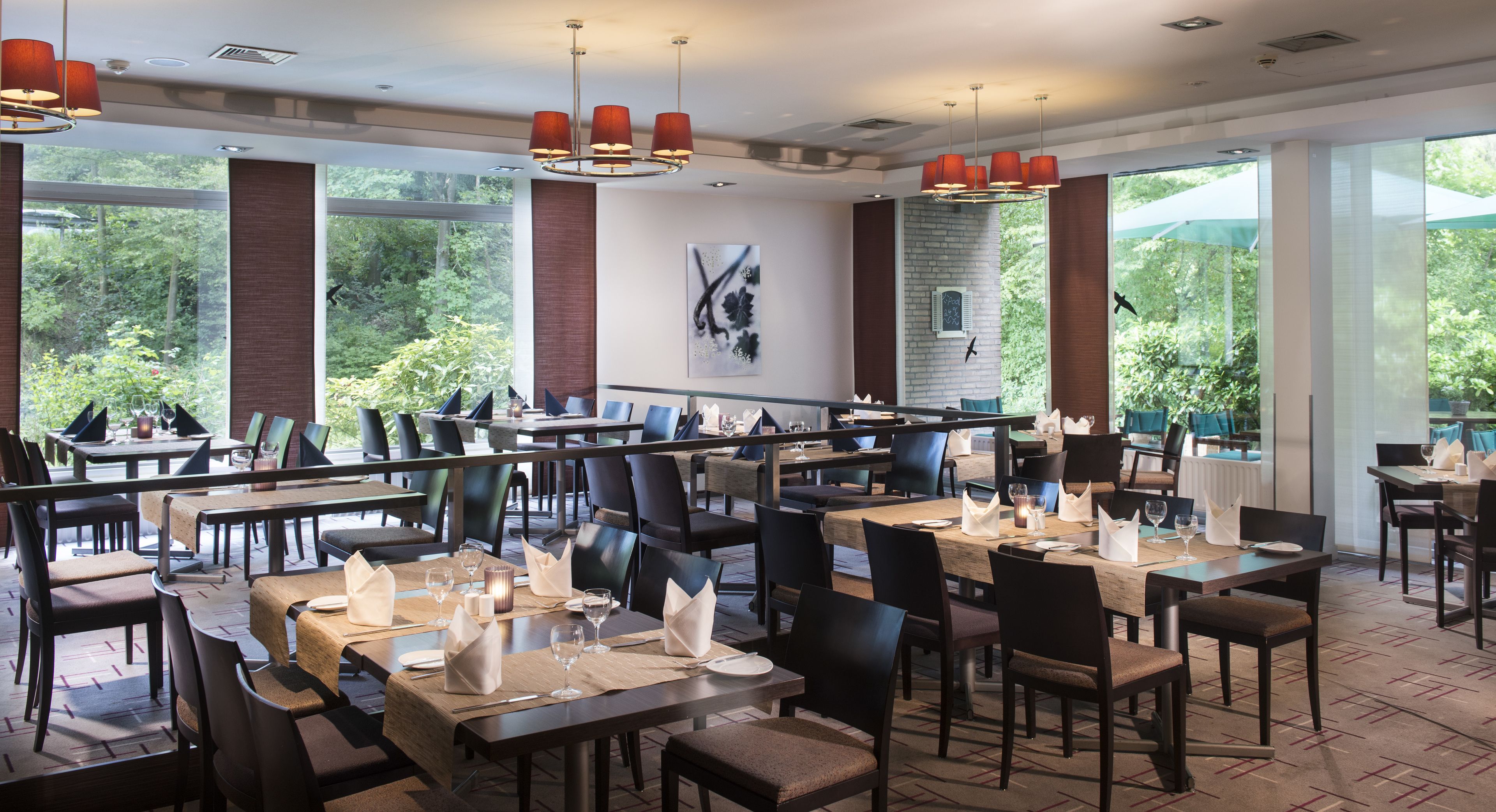A restaurant with multiple tables and chairs, large windows with a view of greenery, and modern pendant lighting.