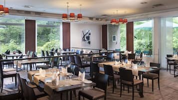 Ein Restaurant mit mehreren Tischen und Stühlen, großen Fenstern mit Blick ins Grüne und moderner Pendelleuchten.