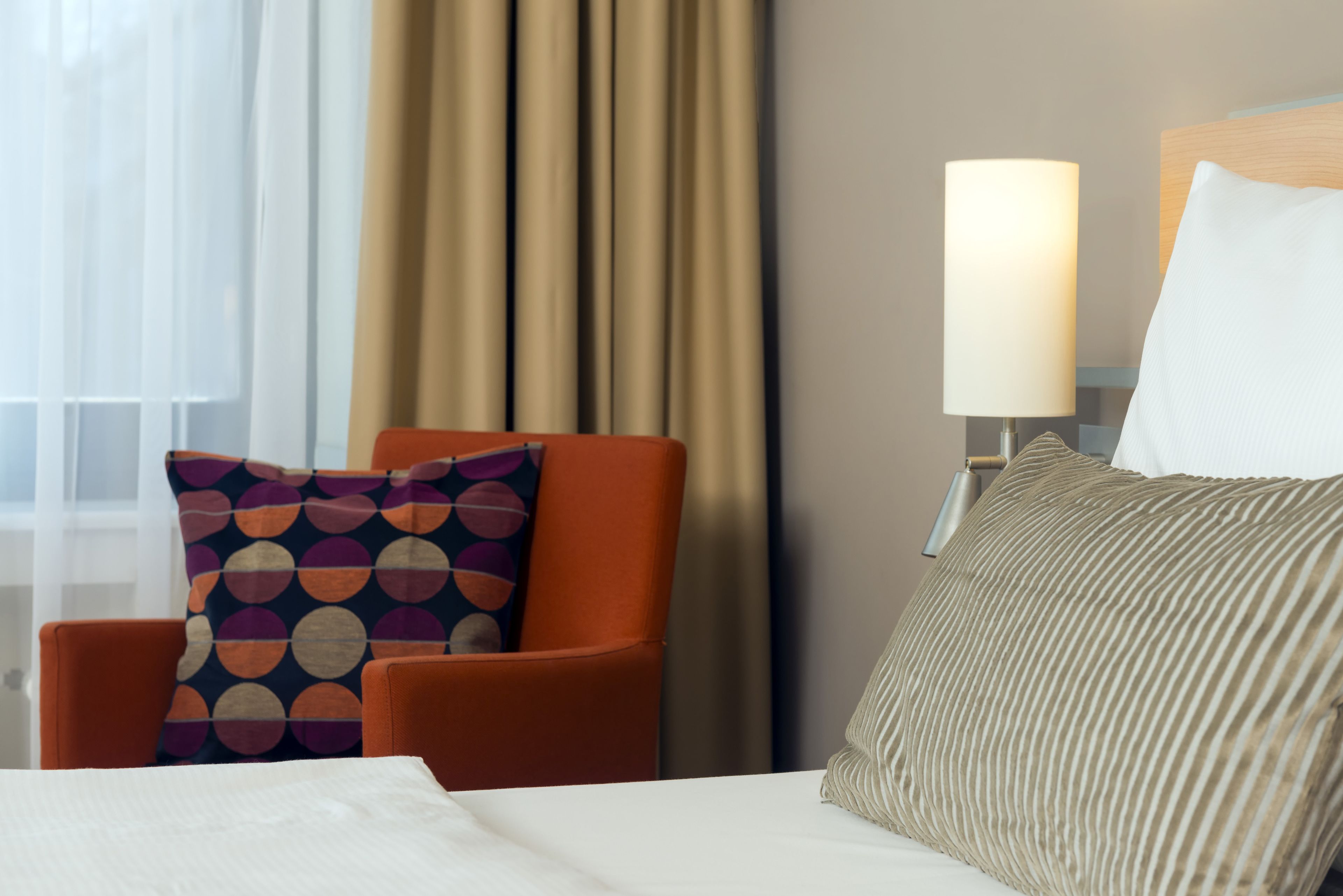 Une chambre d’hôtel avec un lit, une chaise, une lampe et une fenêtre avec des rideaux.
