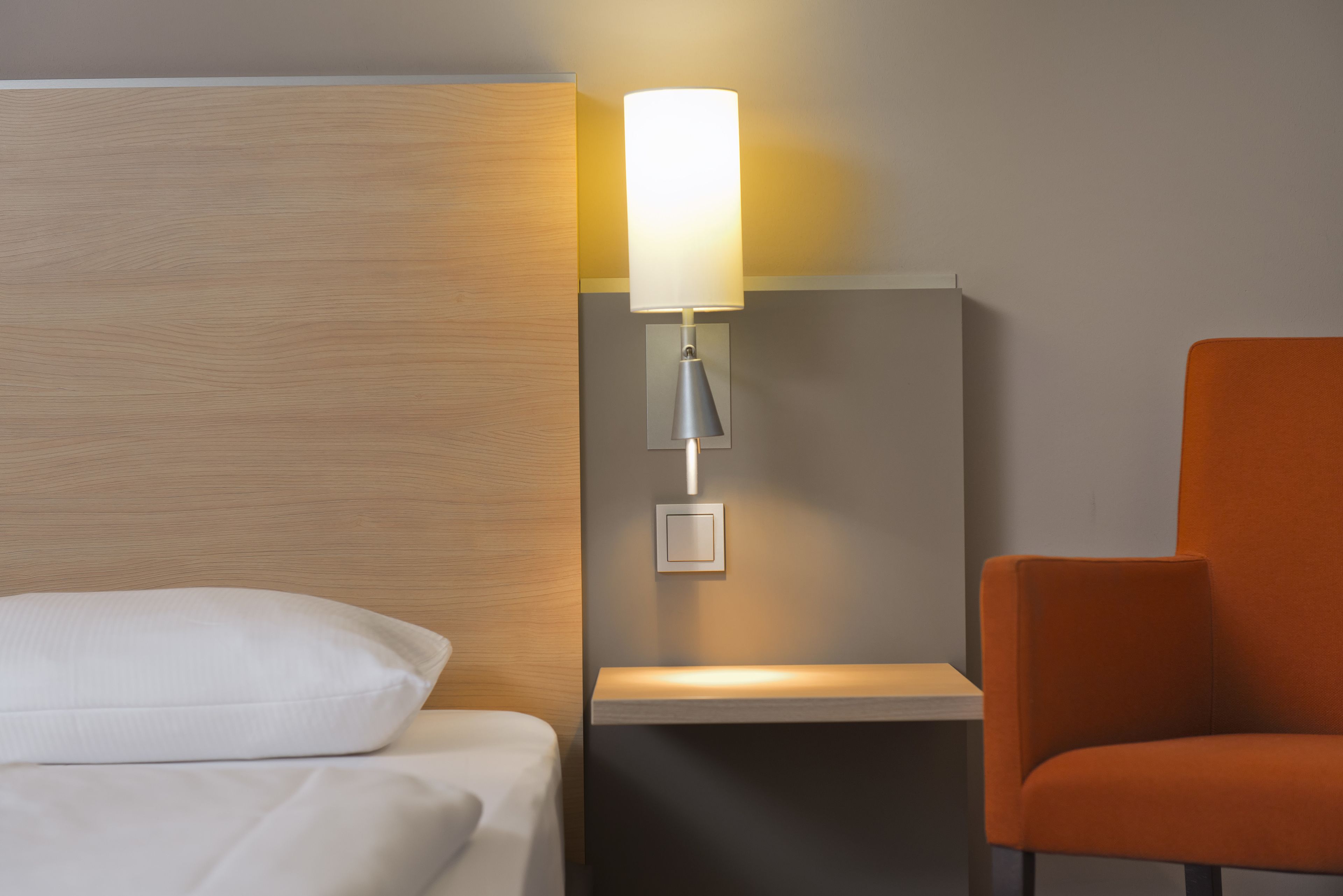 A hotel room with a bed, a nightstand, a lamp, and an orange chair.